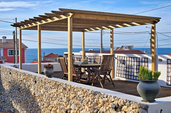 A wooden pergola with a table and chairs on a rooftop terrace with a view of the sea.