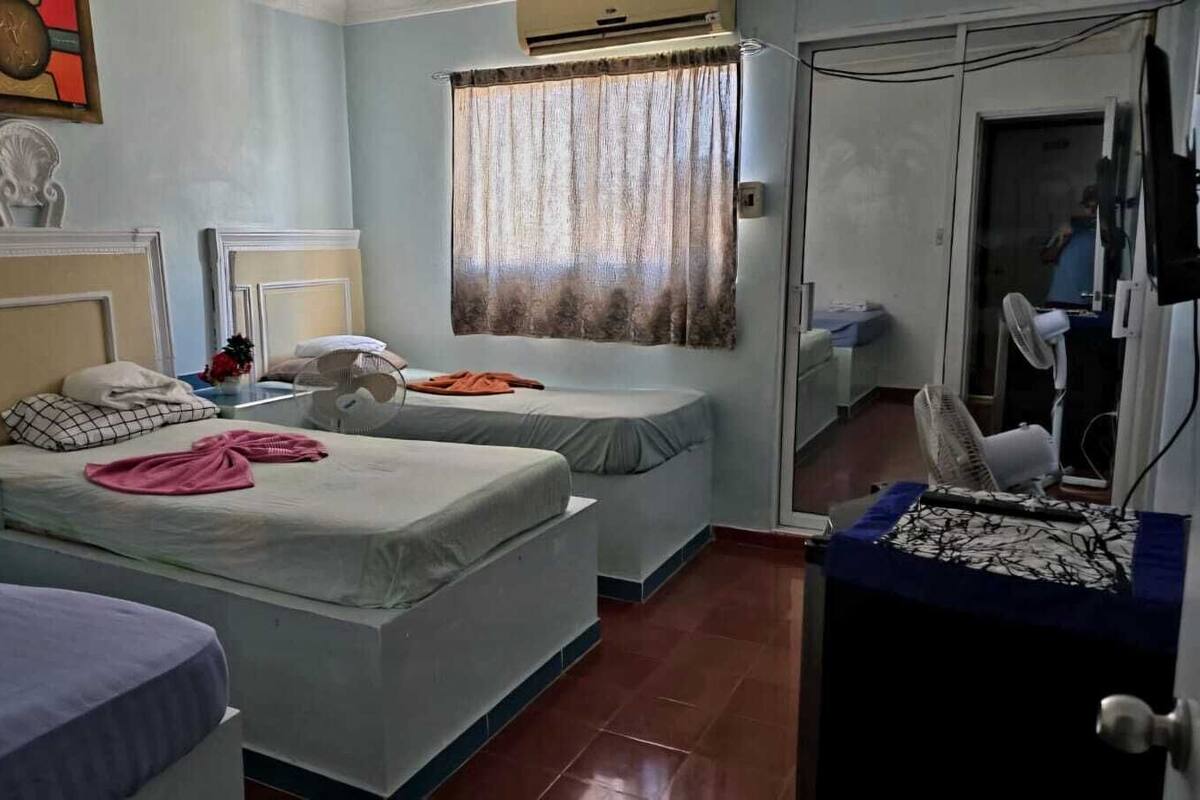 A hotel room with two single beds, a small table with a fan, a TV, and a window with curtains.