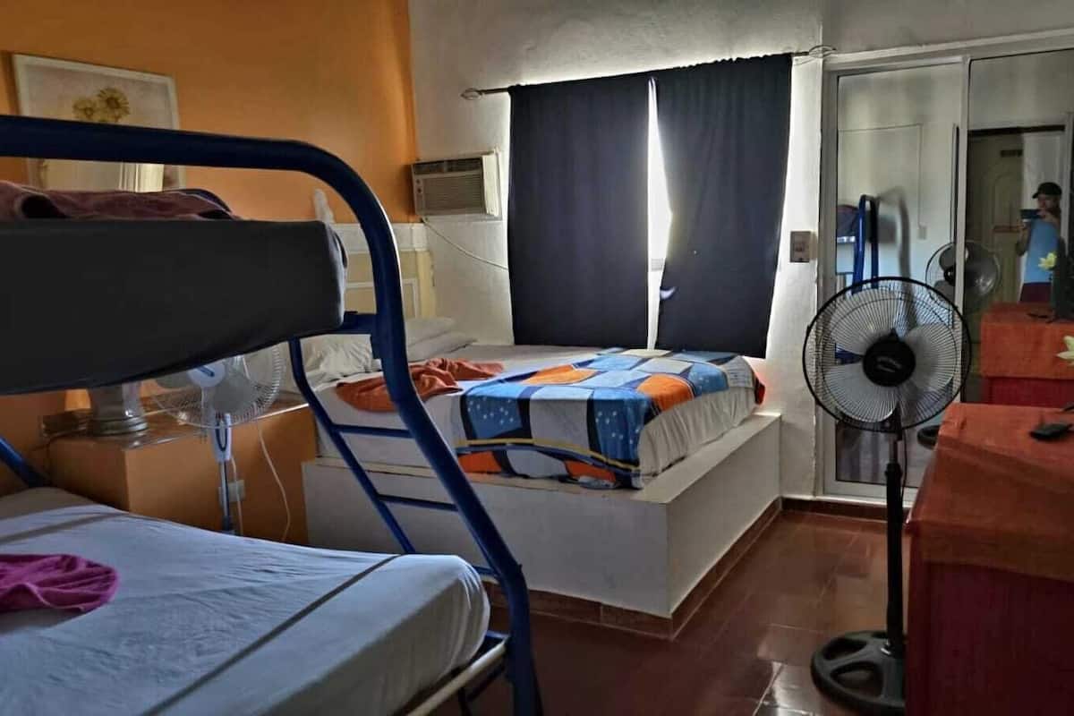A room with bunk beds, a standing fan, a desk with a vase of flowers, and a door leading to another room.