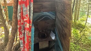 Een klein houten huisje met een enkel houten toilet, een gordijn en uitzicht op het omliggende bos.