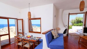 Ein geräumiges Zimmer mit Esstisch, Stühlen, einem blauen Sofa und einem Fernseher. Große Fenster bieten Blick auf die felsige Küste.