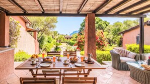 Un espacio comedor al aire libre techado con una mesa preparada para dos, rodeada de sillas, un sofá de mimbre y una mesa con cubierta de vidrio.