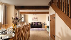 A dining area with a wooden table set for a meal, a sofa in the background, and a staircase with wooden railings. - Webbington Farm Holiday Cottages (Axbridge)