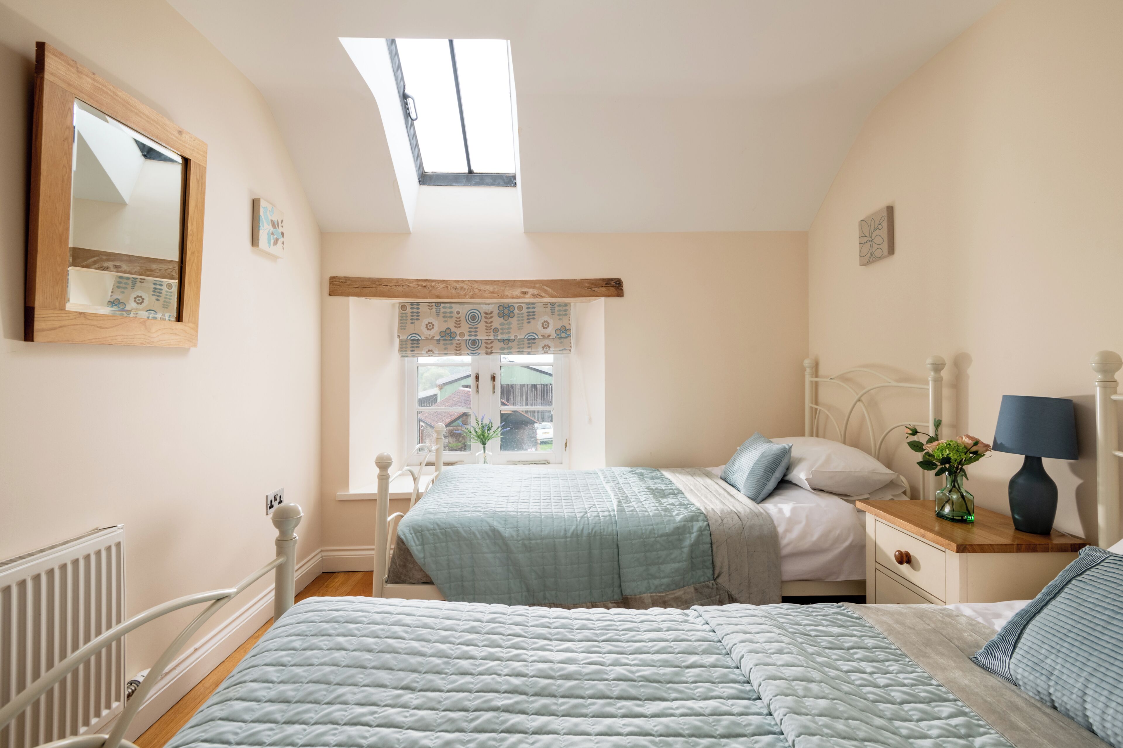 Ein Schlafzimmer mit zwei Betten, einem Oberlicht, einem Fenster mit gemusterten Vorhängen, einem Nachttisch mit Lampe und einem Spiegel an der Wand.