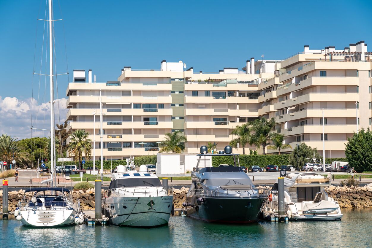 Een jachthaven met verschillende boten afgemeerd, een modern appartementencomplex op de achtergrond, en een heldere blauwe lucht.