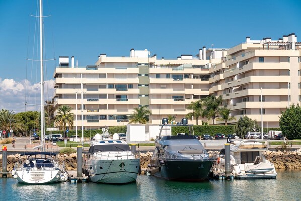 Un puerto deportivo con varias embarcaciones amarradas, un edificio de apartamentos moderno al fondo y un cielo azul despejado.