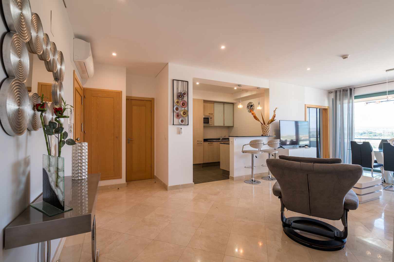 A modern living room with a large window, a kitchen in the background, and a dining area with bar stools.
