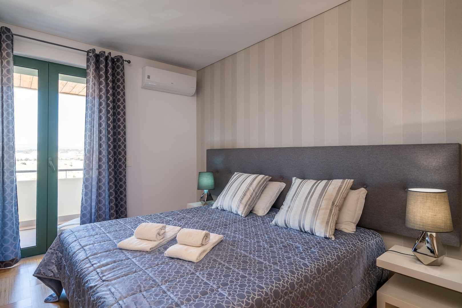 Hotelkamer met een bed, twee kussens, een nachtkastje met lamp, een raam met gordijnen en een airconditioning.