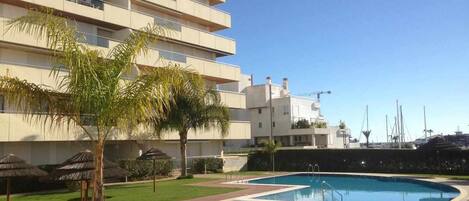 A modern building with a swimming pool, palm trees, and a clear sky.