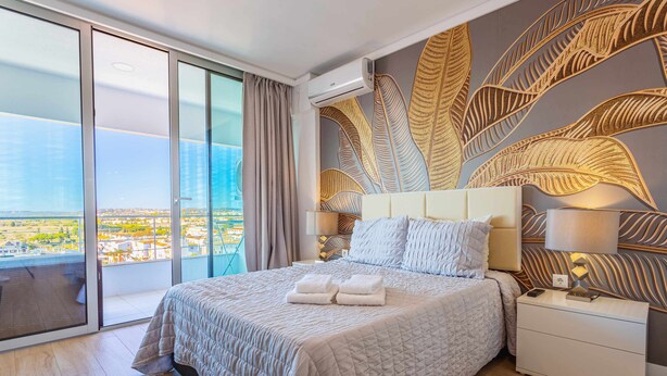 Une chambre d’hôtel moderne dotée d’un grand lit, d’une table de chevet et offrant une vue sur la ville grâce à de grandes fenêtres du sol au plafond.