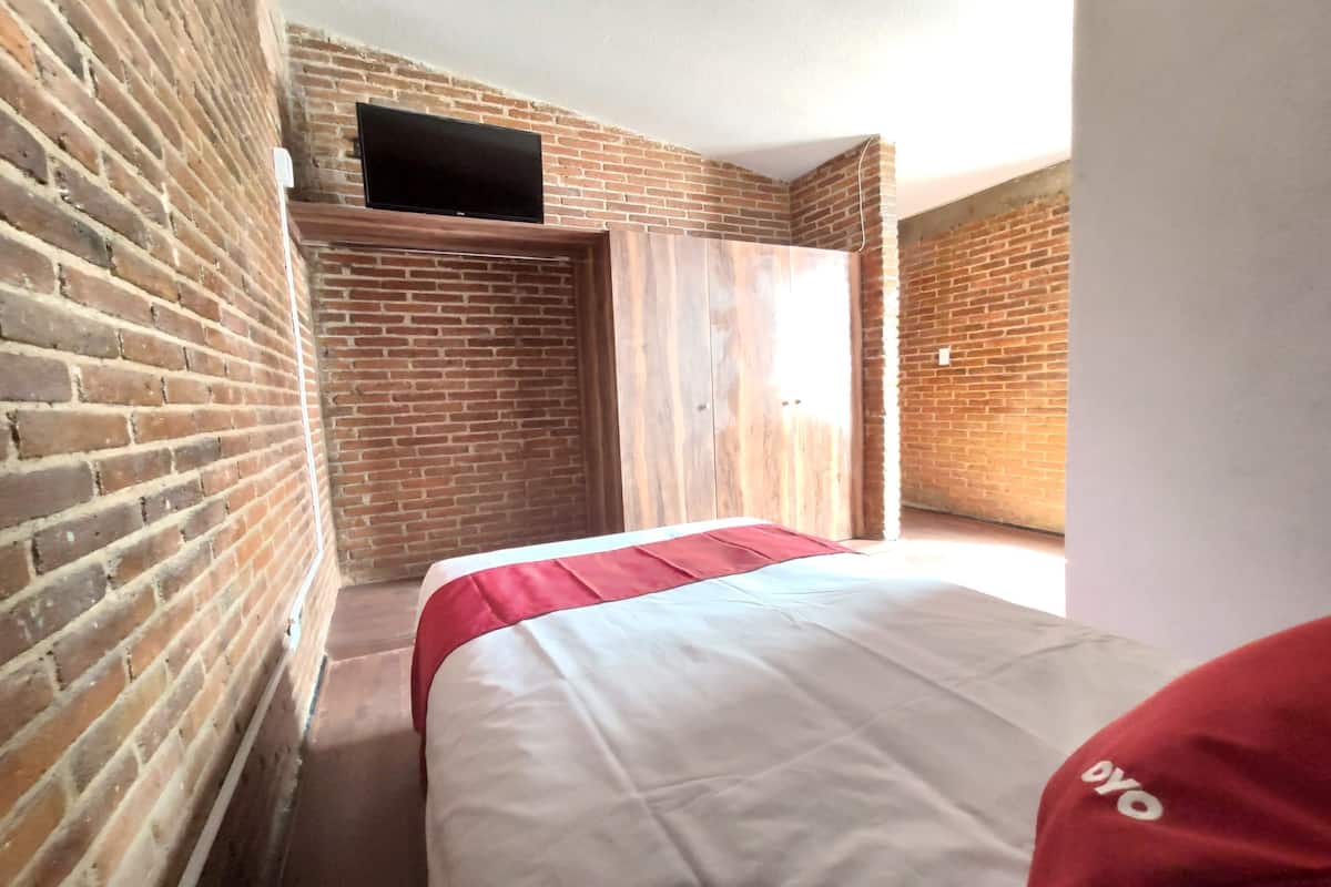 A room with a brick wall, a bed with a red and white bedspread, a television mounted on the wall, and a wooden door.