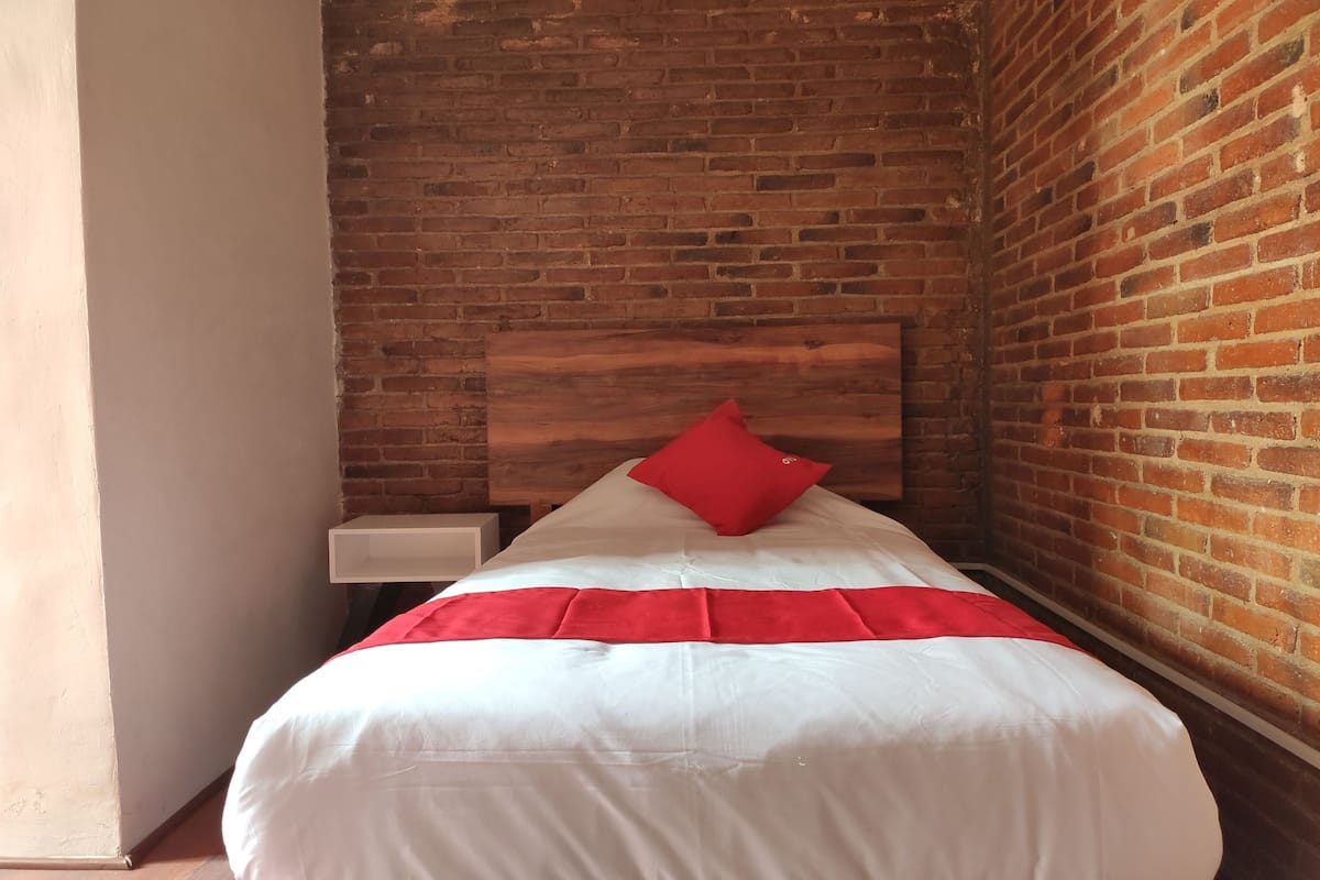 A single bed with white bedding and a red accent pillow in a room with exposed brick walls and a white nightstand.