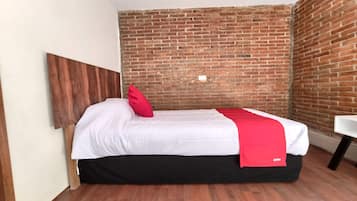 Una cama individual con colchón blanco y falda negra, una almohada roja y una manta roja con el logotipo "OYO". La habitación tiene una pared de ladrillo y suelo de madera.