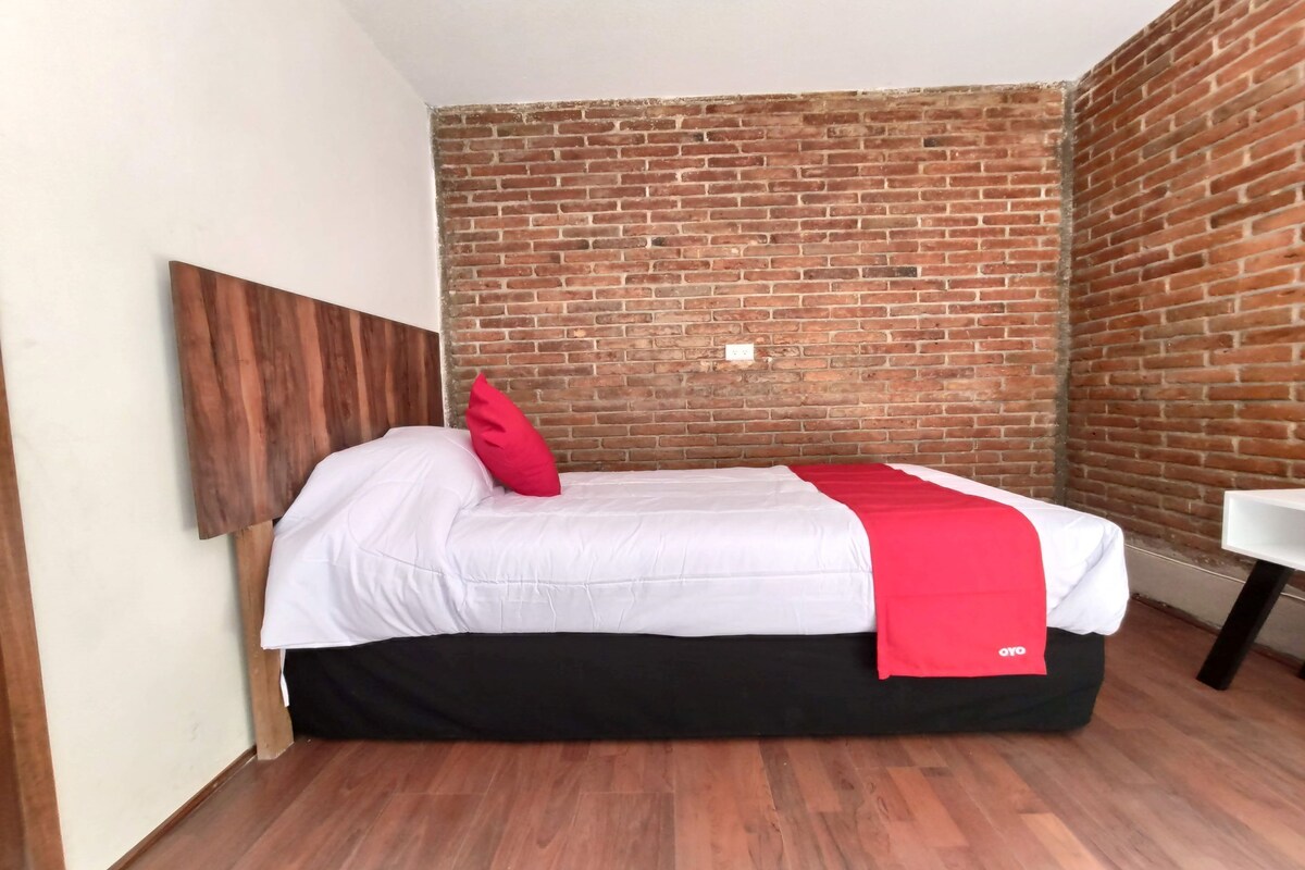 A single bed with a white mattress and black skirt, a red pillow, and a red blanket with 'OYO' branding. The room has a brick wall and wooden flooring.