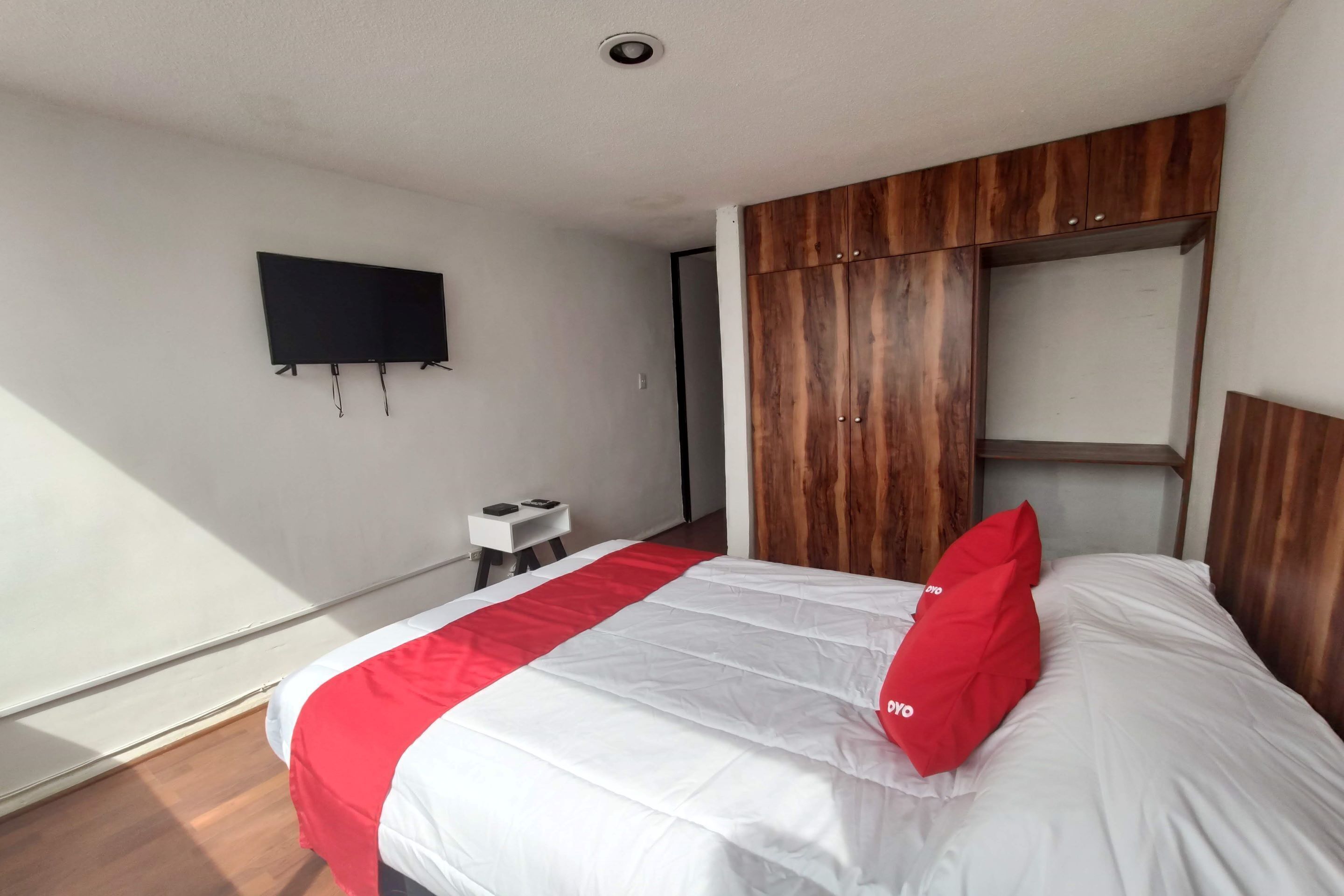 A hotel room with a bed, a TV mounted on the wall, a small table, and a wooden wardrobe.