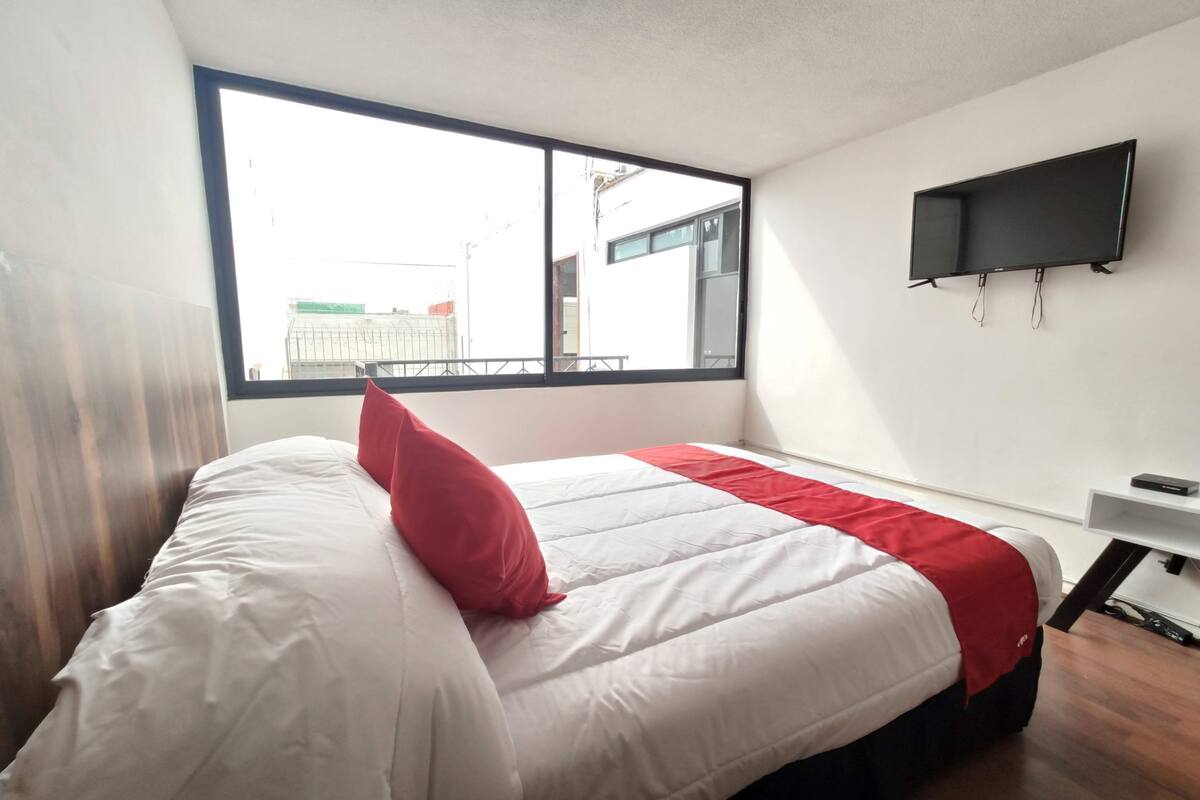 A hotel room with a large window, a bed with white and red bedding, a flat-screen TV mounted on the wall, and a wooden floor.