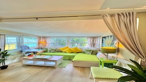 Un salón luminoso con un sofá verde, almohadas amarillas, una mesita de centro y una silla. Hay ventanas con cortinas, una lámpara y una planta.