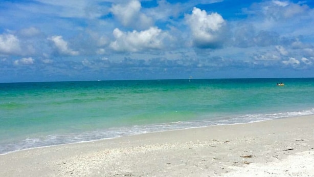 Una playa con agua turquesa cristalina y una orilla arenosa bajo un cielo azul brillante con nubes dispersas.