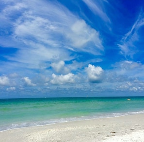Ein Strand mit klarem türkisblauem Wasser und einem sandigen Ufer unter einem strahlend blauen Himmel mit vereinzelten Wolken.