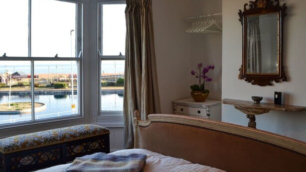 A hotel room with a bed, a bench, a window with a view of a body of water and a building, a mirror, and a small table with a plant. - Georgian Seafront fisherman’s cottage | Sea views | Old Town (Hastings)
