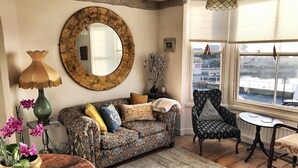 A cozy living room with a patterned sofa, a round mirror, a table with a vase of flowers, and a view of a canal through the window. - Georgian Seafront fisherman’s cottage | Sea views | Old Town (Hastings)