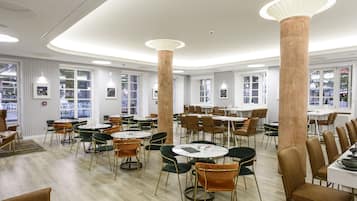 A spacious dining area with round tables and chairs, large columns, and large windows with views.