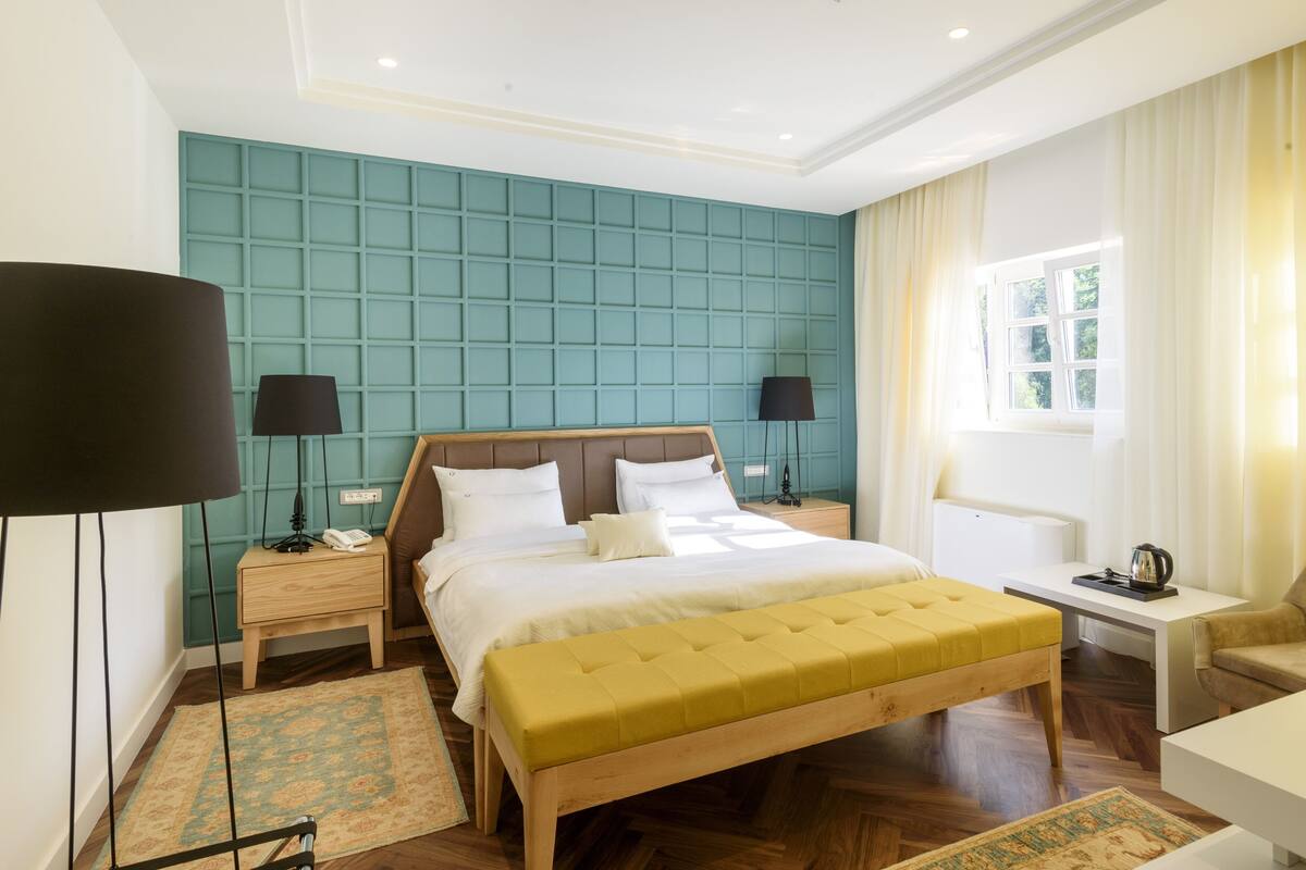 A hotel room with a large bed, two bedside tables, a bedside lamp, a small table with a chair, a window with curtains, and a wooden floor.
