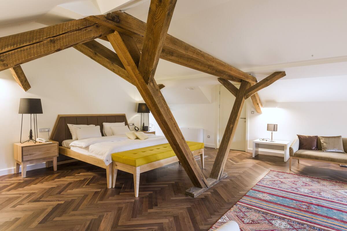 A bedroom with a herringbone floor, a bed with a yellow accent, wooden beams, a bedside table with a lamp, and a sofa with cushions.
