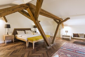 Un dormitorio con suelo de espiga, cama con detalles amarillos, vigas de madera, mesita de noche con lámpara y sofá con cojines.