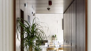 Un pasillo con un árbol en maceta, piso de madera, una zona de comedor con mesa y sillas, y una cocina al fondo.