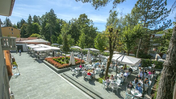 Un área de café al aire libre con mesas y sillas, rodeada de árboles y edificios.