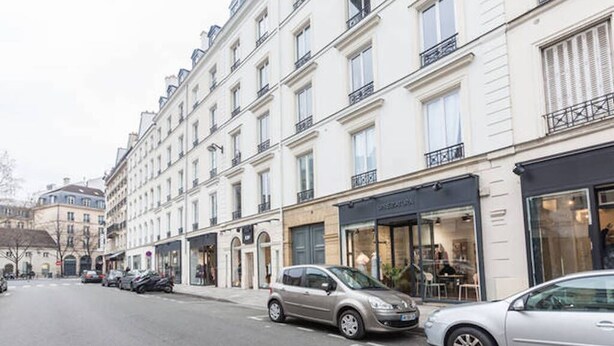 A street view of a row of white buildings with storefronts, parked cars, and a clear sky. - Apartment Paris Center "Marais" - 4 people (Paris)