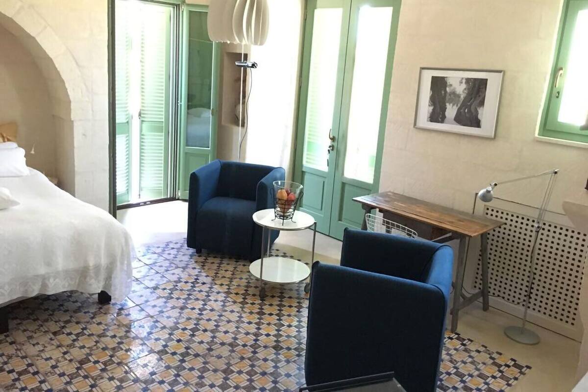 A room with a bed, two blue armchairs, a small round table with a fruit basket, a desk with a lamp, and a radiator.