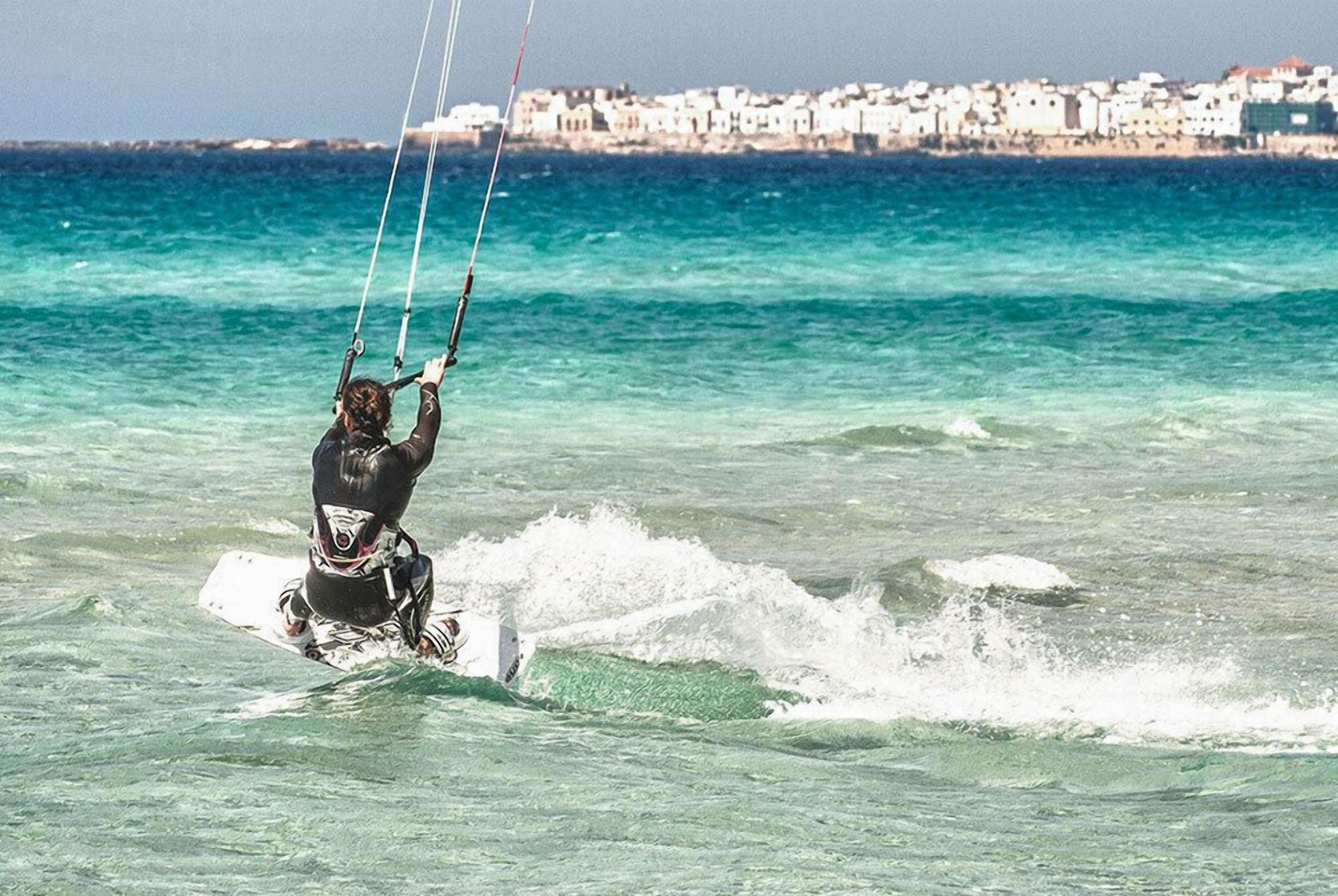 Una persona practica windsurf en un día despejado, con un pueblo costero al fondo.
