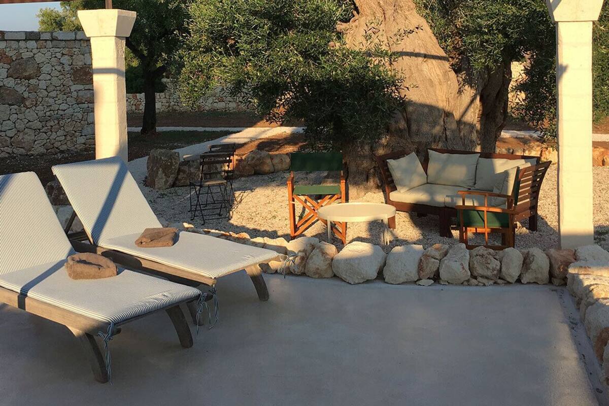 A sunlit outdoor seating area with lounge chairs and a small table, surrounded by a stone wall and a large tree.