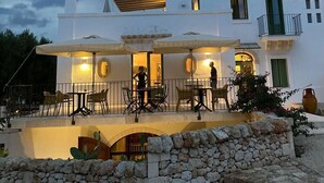 A white building with a stone wall in front, outdoor seating with umbrellas, and a balcony with a railing. - Masseria Sottomasi (Fasano)