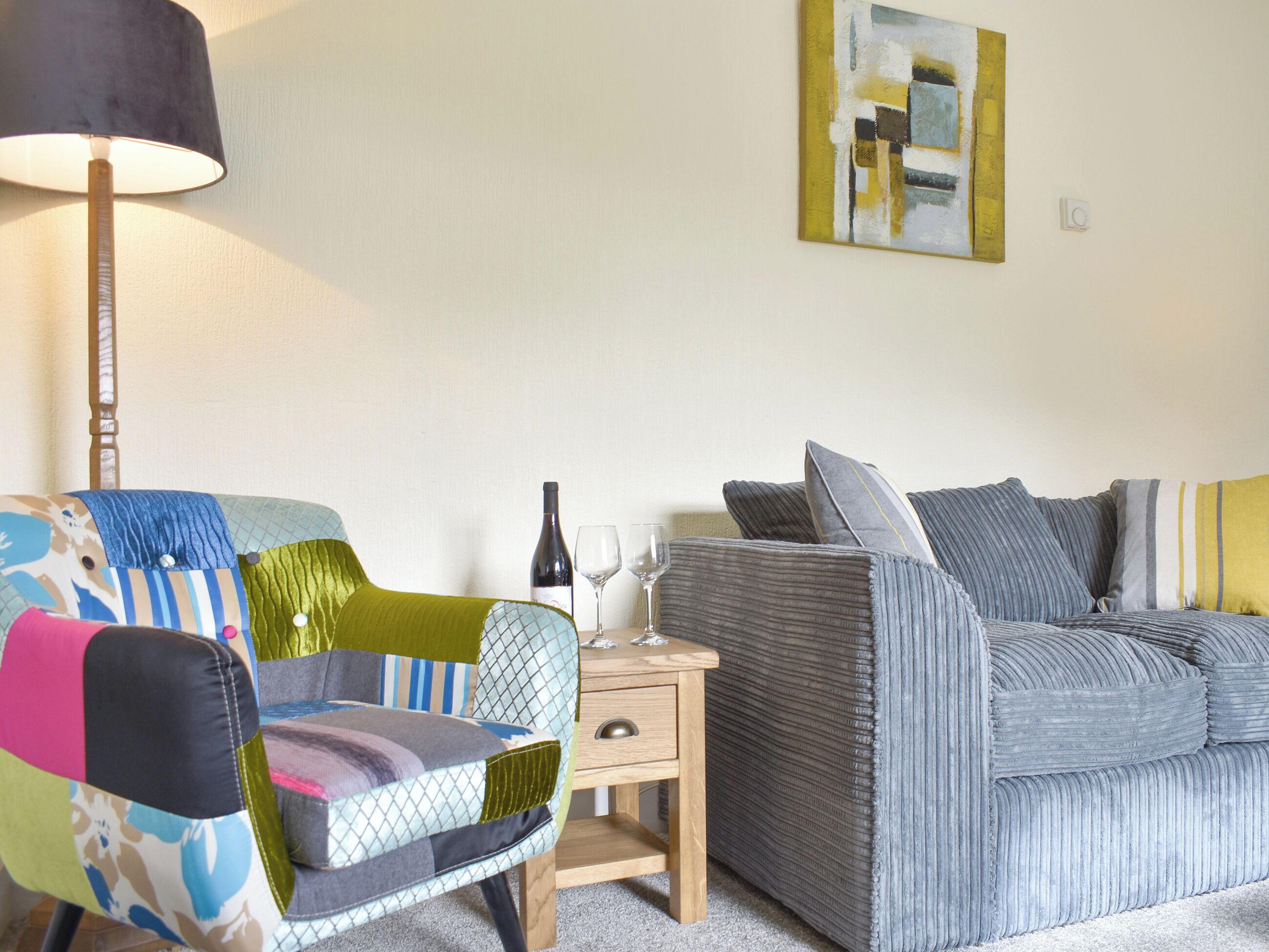 A room with a colorful armchair, a grey sofa, a wooden nightstand, and a lamp.