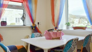 Un coin repas avec une table blanche, des chaises colorées et une vue sur une maison par la fenêtre.