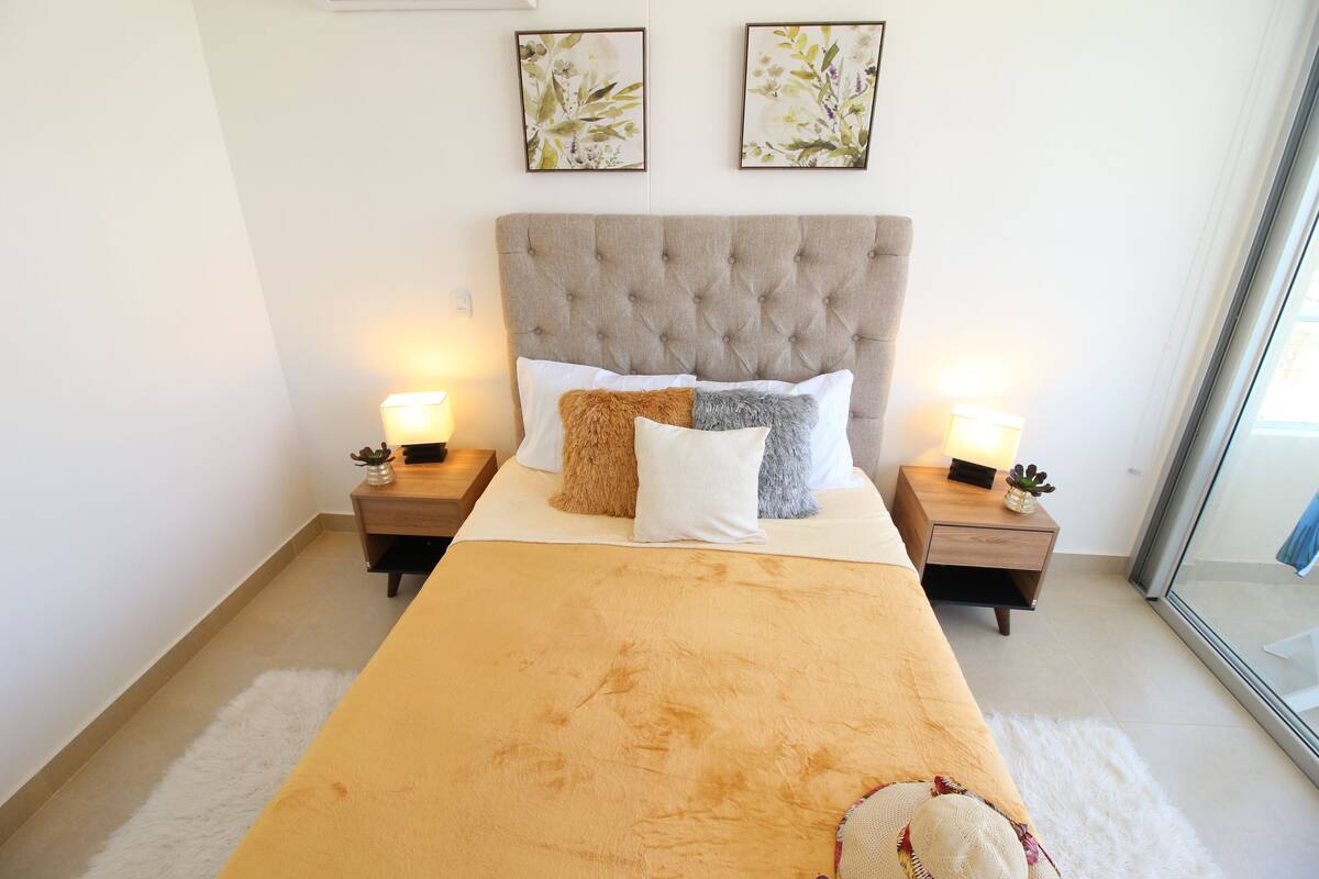 A neatly made bed with a beige headboard, two bedside tables with lamps, and two framed floral artworks on the wall.