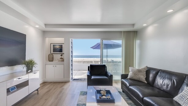 A modern living room with a large flat-screen TV, a leather sofa, and a view of the beach through large sliding glass doors. - Remodeled Beachfront Mission Beach Condo on the boardwalk with Amazing Views! (San Diego)