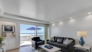 A modern living room with a black leather sofa, a coffee table, and a lamp. There is a view of the beach through a glass door. - Remodeled Beachfront Mission Beach Condo on the boardwalk with Amazing Views! (San Diego)