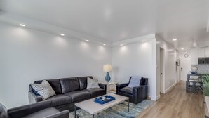 A modern living room with a sofa, chairs, a coffee table, and a lamp. There is a kitchen area visible in the background. - Remodeled Beachfront Mission Beach Condo on the boardwalk with Amazing Views! (San Diego)