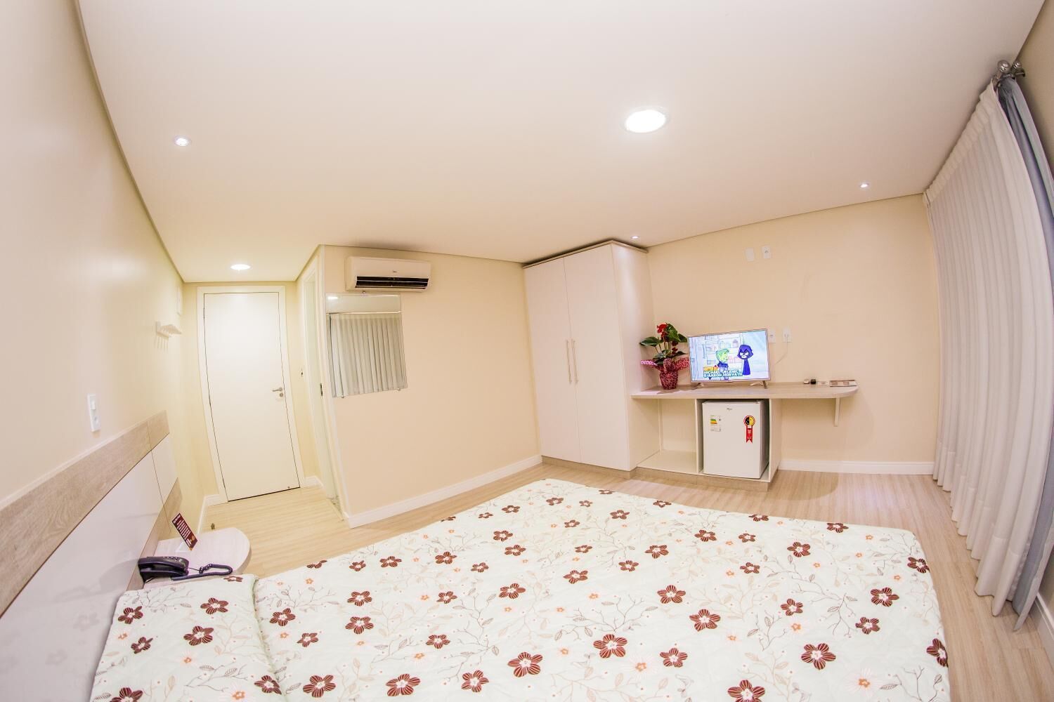 Quarto com cama, TV, mini-frigorífico e janela com cortinas.