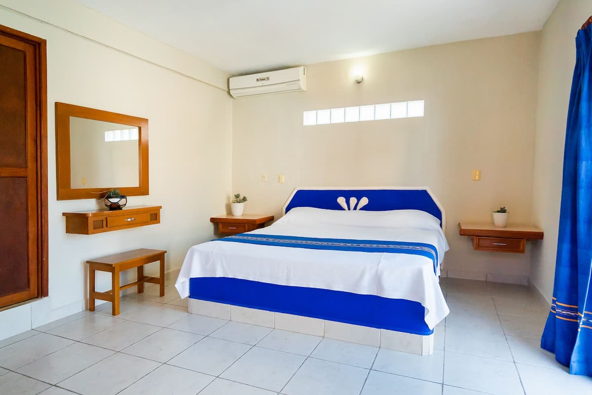 A bedroom with a bed, a wooden bench, a mirror, a wall-mounted shelf, and an air conditioning unit.