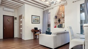 A modern living room with a white sofa, a white armchair, and a wooden floor. There is a bookshelf with books and decorative items, a small table with a laptop, and a door with a heart-shaped decoration. - SARA'S LUXURY HOME 5 minutes from St Mark's Square (Venice)