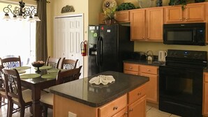 Une cuisine avec des meubles de rangement en bois, un réfrigérateur noir, un four à micro-ondes et une cuisinière. On y trouve une table à manger avec des chaises et un lustre.