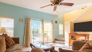 A living room with a ceiling fan, a glass coffee table, and a TV on a wooden stand. - Private Screened Splash Pool Overlooking Tranquil Lake Right Beside Disney World by Florida Dream Homes (Kissimmee)