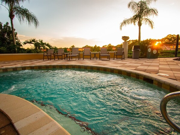 A swimming pool with a tiled edge, surrounded by lounge chairs and palm trees, with the sun setting in the background.