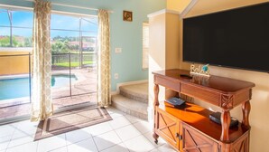 A living room with a TV, wooden cabinet, and a view of a swimming pool. - Private Screened Splash Pool Overlooking Tranquil Lake Right Beside Disney World by Florida Dream Homes (Kissimmee)