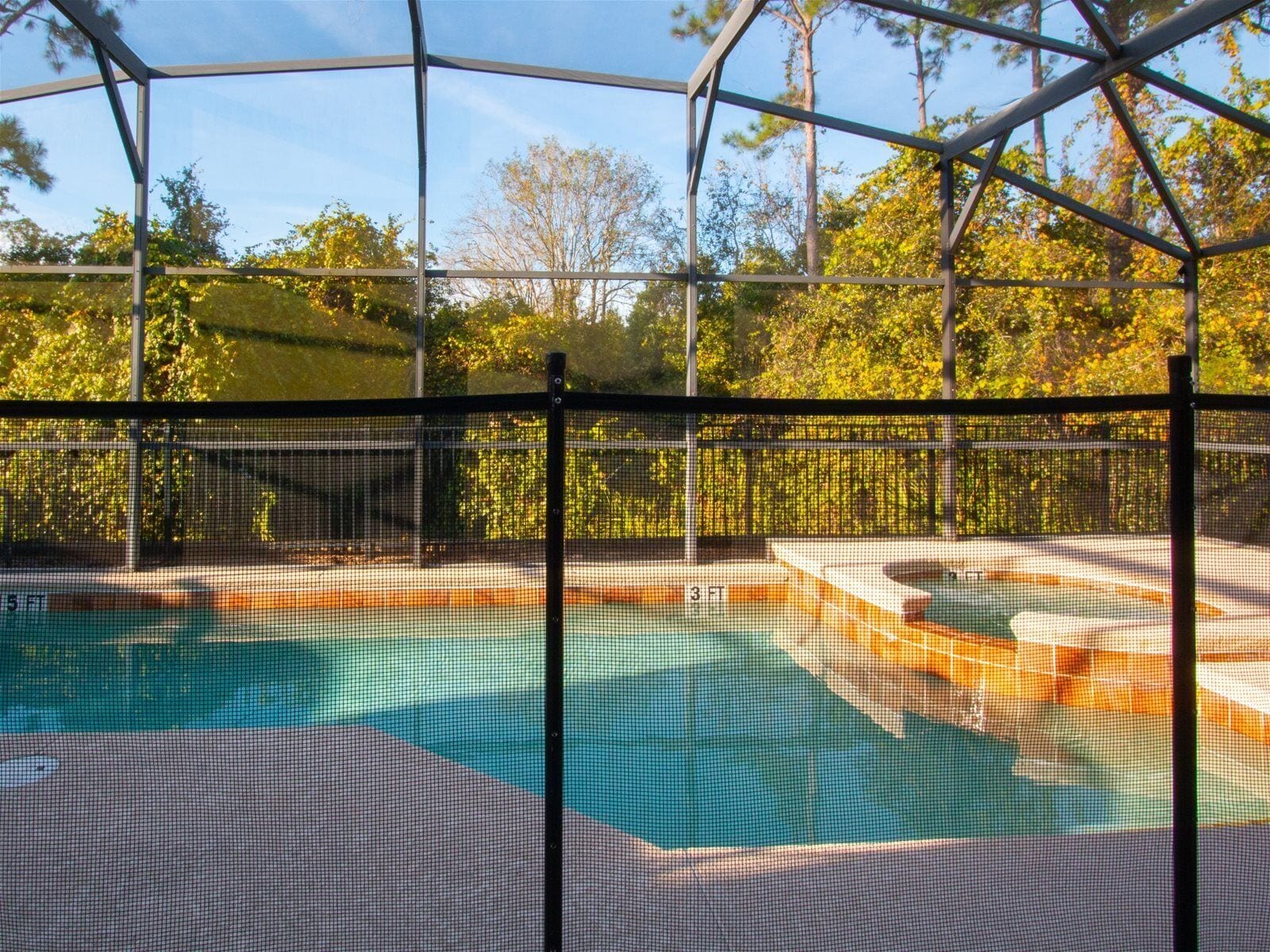A swimming pool with a safety fence, surrounded by trees and a clear sky.